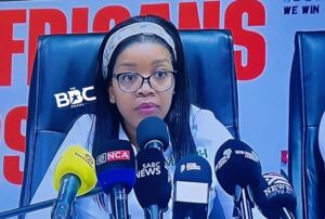 Woman wearing glasses at a press conference, speaking into multiple microphones labeled with news outlets (SABC News, NCA), with a red backdrop behind him/her.