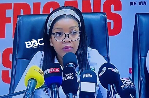 Woman wearing glasses at a press conference, speaking into multiple microphones labeled with news outlets (SABC News, NCA), with a red backdrop behind him/her.