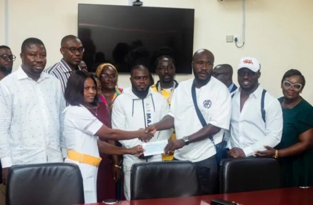 Group of people in a meeting room shake hands as a certificate is handed over in front of a large screen TV on the wall.