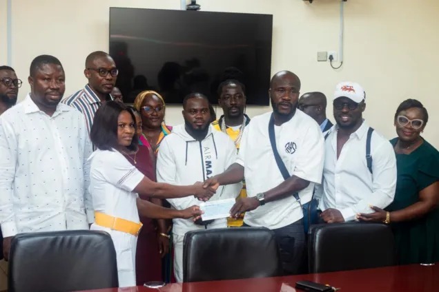 Group of people in a meeting room shake hands as a certificate is handed over in front of a large screen TV on the wall.