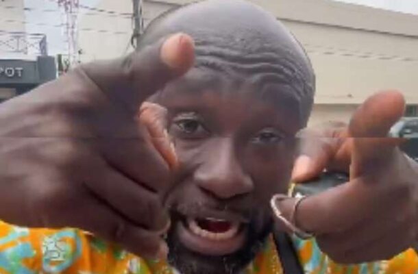 Close-up of a man with a shaved head in a bright orange and teal patterned shirt, pointing at the camera with both hands while speaking, outdoors in an urban area.