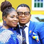 Couple posing together outdoors, the woman in blue lace and braided hair, the man in a blue suit with glasses and patterned tie.
