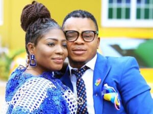 Couple posing together outdoors, the woman in blue lace and braided hair, the man in a blue suit with glasses and patterned tie.