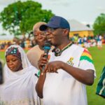 Man in a white sports shirt and cap speaks into a microphone on a football field, with a crowd in the background.
