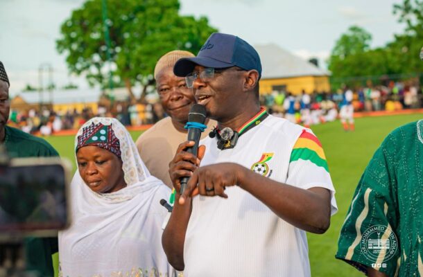 IMG-20260608-WA0001 | Ghanaguardian.com Man in a white sports shirt and cap speaks into a microphone on a football field, with a crowd in the background.