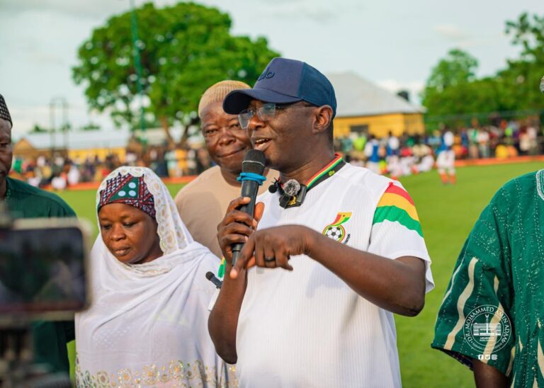 Man in a white sports shirt and cap speaks into a microphone on a football field, with a crowd in the background.