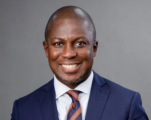 Professional headshot: smiling man in a navy suit and striped tie against a gray background, suitable for a corporate profile.