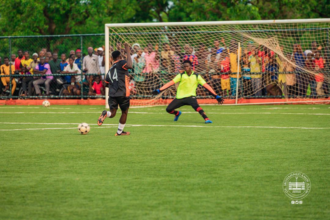 IMG-20260608-WA0005 | Ghanaguardian.com Soccer match action with a player in black kicking toward the goal as a goalkeeper in neon green faces him near the net; a crowd watches from behind the goal on a sunny day.