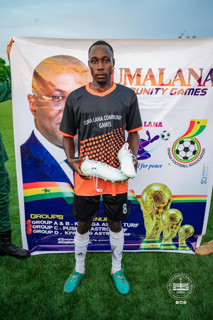 IMG-20260608-WA0010 | Ghanaguardian.com Young male footballer in orange-black kit holds white cleats in front of a banner for Tumala Lana Community Games on a sunny field