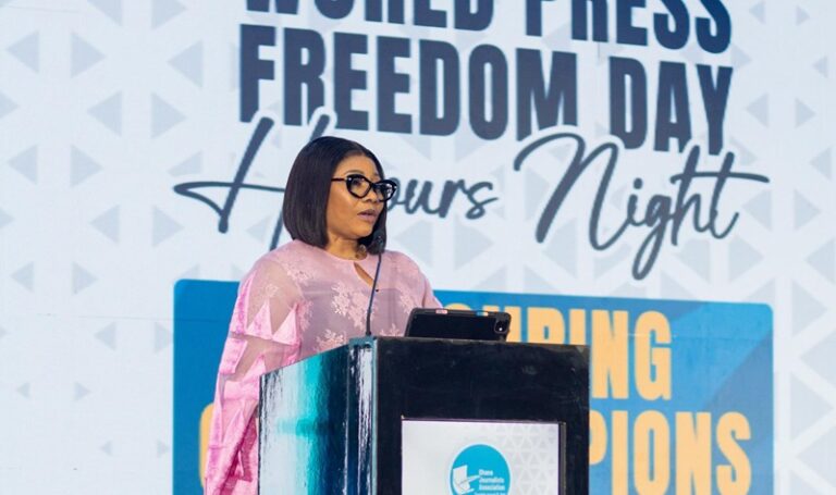 Woman speaker at a podium during World Press Freedom Day event, wearing a pink top and sunglasses, delivering remarks.