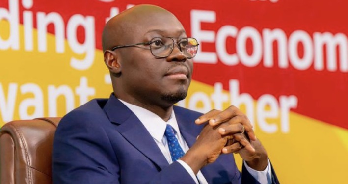 Man in a navy suit and glasses sits on a brown chair with hands clasped, appearing thoughtful at a conference backdrop of red and yellow banners.
