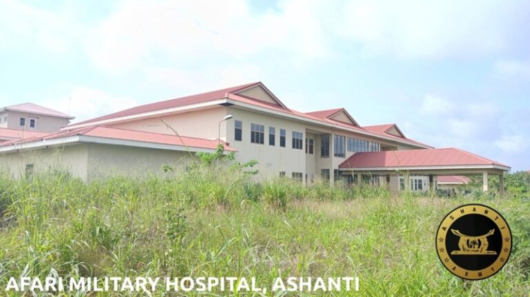 Exterior of Afari Military Hospital in Ashanti: beige building with red roof, set behind tall grass, with a circular emblem on the right.