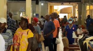 Crowd of people standing in a long queue inside a busy hall, some holding bags, with a nurse in blue uniform nearby.