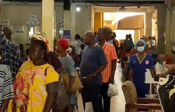 Crowd of people standing in a long queue inside a busy hall, some holding bags, with a nurse in blue uniform nearby.