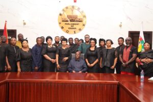 Group of officials in black attire posing for a formal photo in Parliament of Ghana; seated man center, gold seal behind, Ghana flag to the right.
