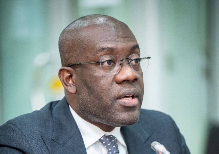 Portrait of a Black man in a navy suit and glasses speaking into a microphone at a formal event.