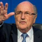 Older man wearing glasses, a dark suit and blue tie, speaking at a press conference with his hand raised in a gesture against a blue backdrop.