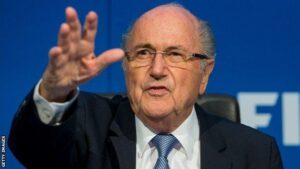 Older man wearing glasses, a dark suit and blue tie, speaking at a press conference with his hand raised in a gesture against a blue backdrop.