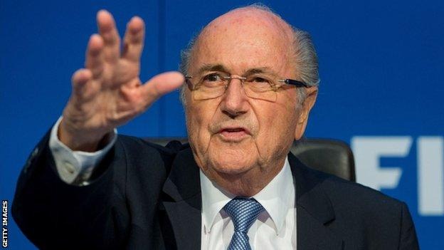 Older man wearing glasses, a dark suit and blue tie, speaking at a press conference with his hand raised in a gesture against a blue backdrop.