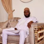 Man in white traditional attire sits on an ornate beige sofa in a luxurious living room, looking at the camera with a relaxed pose and a folder on his lap-to his sideoiled closet?