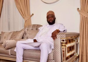 Man in white traditional attire sits on an ornate beige sofa in a luxurious living room, looking at the camera with a relaxed pose and a folder on his lap-to his sideoiled closet?