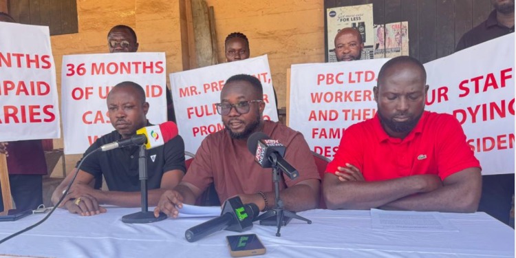 Panelists sit at a press conference table with microphones as protesters hold red-on-white signs about unpaid salaries in the background, conveying a wages protest theme.