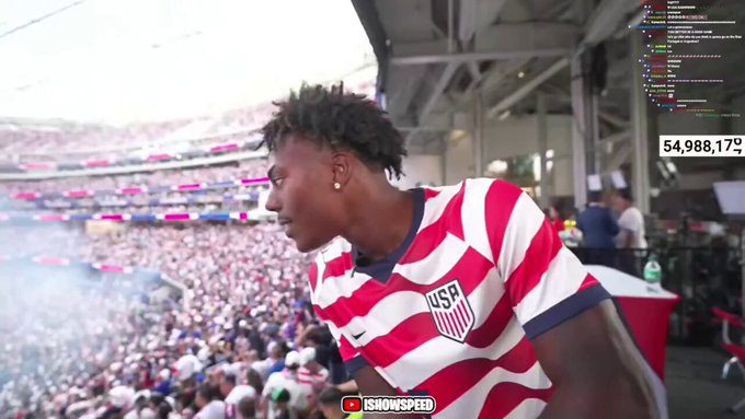 Man in a red‑white USA striped jersey leans over a railing at a packed stadium, crowd cheering behind him.