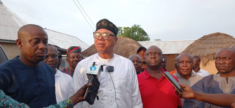Man in white traditional attire and a cap speaks to reporters with microphones outdoors.