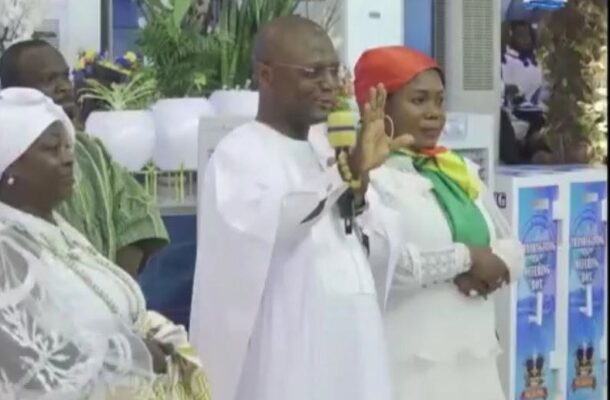 Man in white traditional attire speaks into a microphone with his hand raised, standing beside a woman in a red headscarf at a formal event.