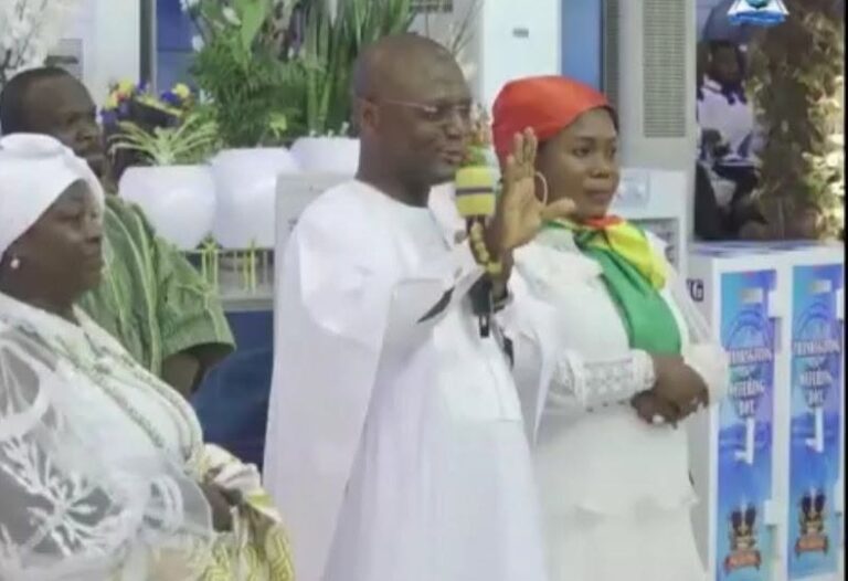 Man in white traditional attire speaks into a microphone with his hand raised, standing beside a woman in a red headscarf at a formal event.
