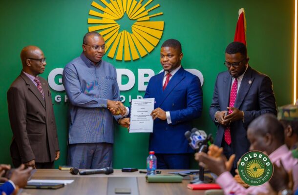 Four men in suits at a press conference; two center men shake hands and hold a document against a green wall?