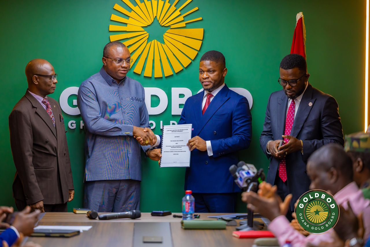 Four men in suits at a press conference; two center men shake hands and hold a document against a green wall?