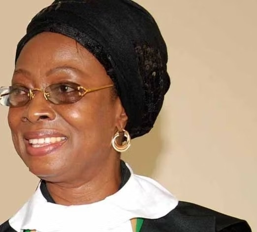 Smiling woman wearing a black head wrap, gold-rimmed glasses, and gold hoop earrings against a beige background.