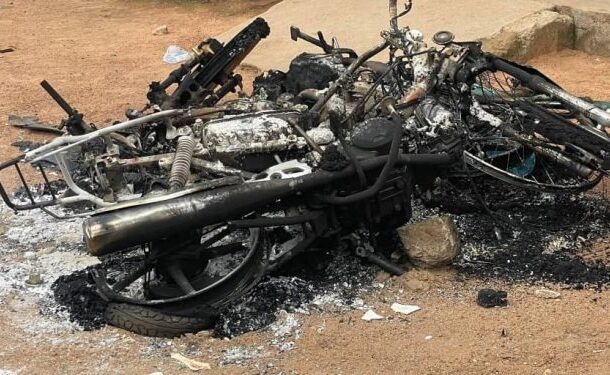 Burned-out motorcycle frame and charred debris on dusty ground in an outdoor setting.