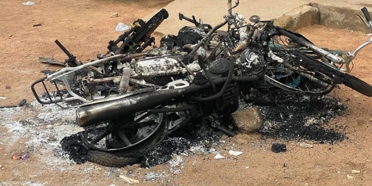Burned-out motorcycle frame and charred debris on dusty ground in an outdoor setting.