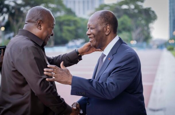 Two men greet each other with a warm handshake and smiles outdoors, one in a dark shirt and the other in a navy suit.