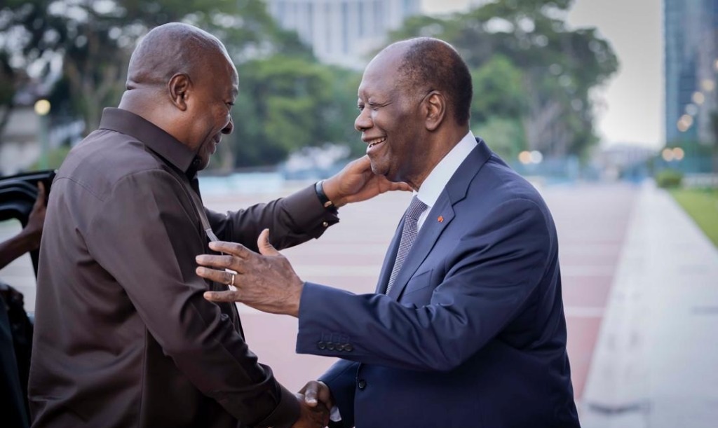 Two men greet each other with a warm handshake and smiles outdoors, one in a dark shirt and the other in a navy suit.