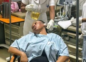 Male patient in a hospital gown reclining on a bed as a clinician pours yellow liquid into his mouth from a pitcher for treatment.