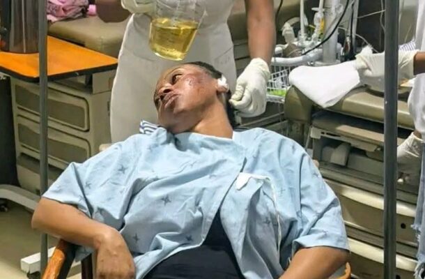 Patient in a hospital gown lying back as a nurse pours a yellow liquid into his mouth in a clinical room with medical equipment nearby.