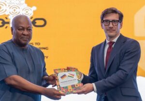 Two men in formal attire exchange a colorful booklet with flag images on a bright yellow backdrop.