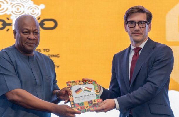 Two men in formal attire exchange a colorful booklet with flag images on a bright yellow backdrop.