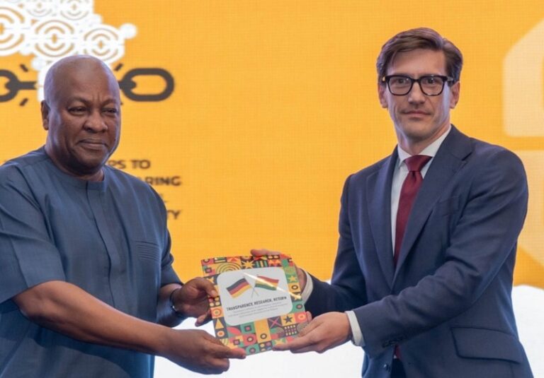 Two men in formal attire exchange a colorful booklet with flag images on a bright yellow backdrop.