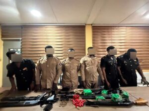 Eight uniformed personnel stand behind a table with seized rifles, ammunition, and green boxes on a room table; faces blurred for privacy.