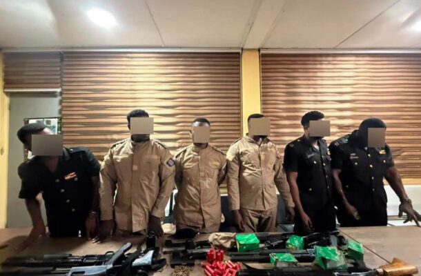 IMG-20260621-WA0068 | Ghanaguardian.com Eight uniformed personnel stand behind a table with seized rifles, ammunition, and green boxes on a room table; faces blurred for privacy.