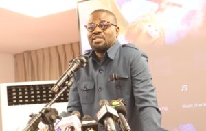 Man in a blue shirt and round glasses speaks into multiple microphones at a press event, with a projector screen behind him.