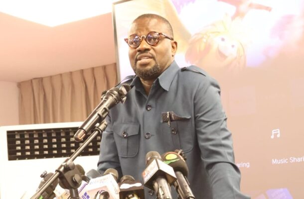 Man in a blue shirt and round glasses speaks into multiple microphones at a press event, with a projector screen behind him.