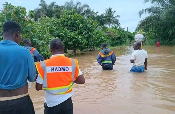 IMG-20260623-WA0099 | Ghanaguardian.com Group of people wading through waist-deep floodwater; one wears an orange NADMO vest, others in safety gear, with trees in the background.