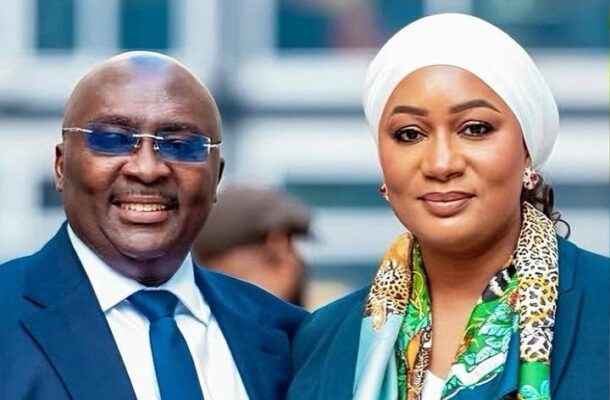 Two well-dressed individuals posing for a photo outdoors: a man in a blue suit and tie with blue sunglasses and a woman wearing a white headscarf and colorful scarf beside him.