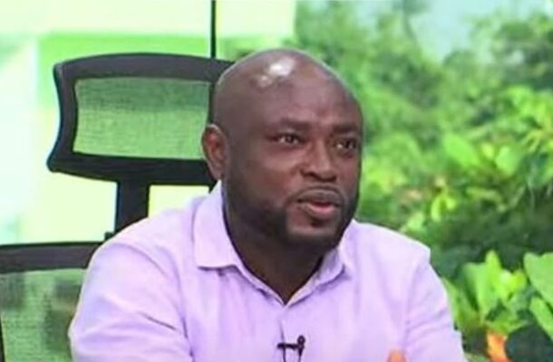 Man in a pink shirt speaking in a studio, with a lapel mic clipped to his shirt