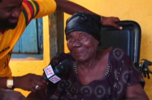 A man interviews an elderly woman in a wheelchair, holding a microphone to her as she sits indoors against a yellow wall.